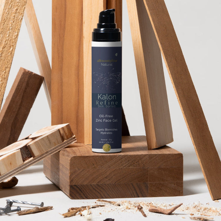 A bottle of Kalon Refine stands on a wooden block in a styled studio setup with wood scraps, screws, and unfinished wood pieces against a light gray background.
