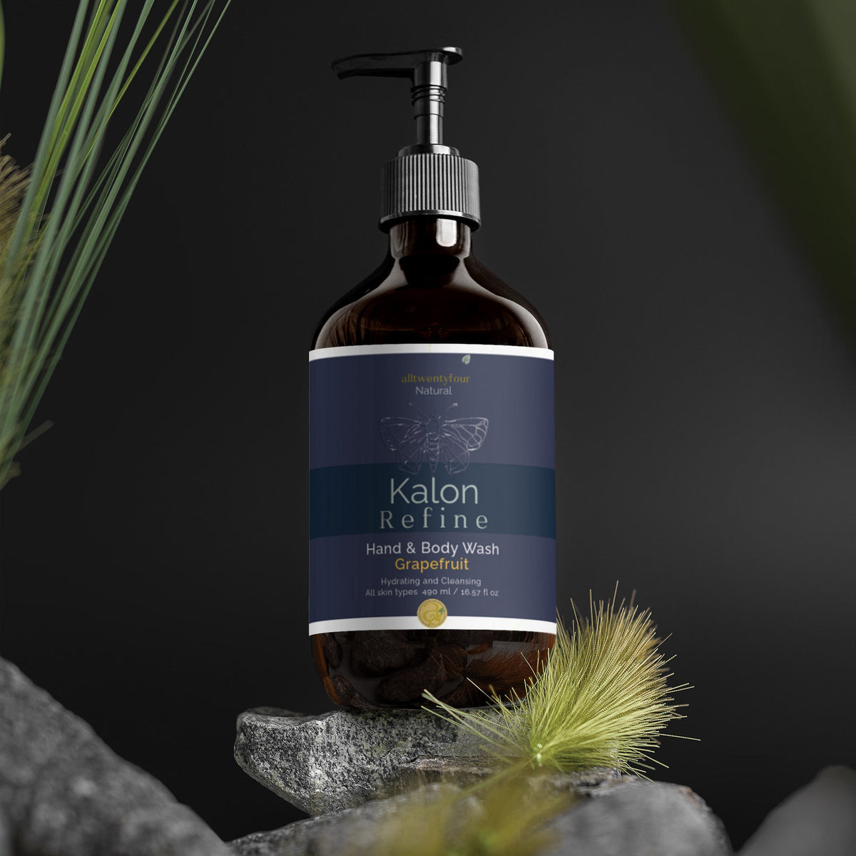 A bottle of Kalon hand and body wash, grapefruit scent, displayed on a stack of gray stones with dried grass against a dark background.