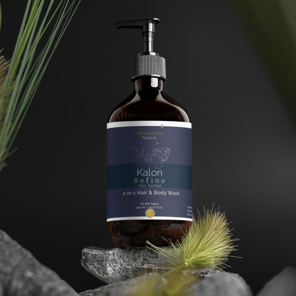 A pump bottle of Kalon 2-in-1 Hair & Body Wash, made of dark amber glass with a navy blue label, styled on a small stone pedestal with blurred natural grass and rocks against a dark background.